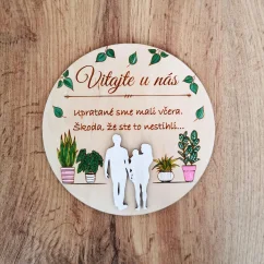 Drevená okrúhla tabuľka s textom "Vitajte u nás, upratané sme mali včera, škoda že ste to nestihli", so siluetami postáv a s ručne maľovanými izbovými rastlinami. 