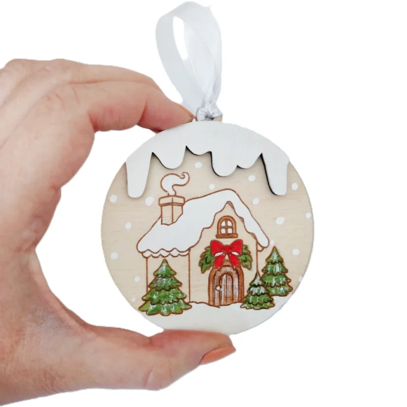 Drevená ozdoba na stromček s gravírovaným a ručne maľovaným zasneženým domčekom s ihličnatými stromčekmi.