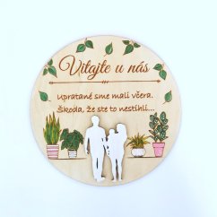 Drevená okrúhla tabuľka s textom "Vitajte u nás, upratané sme mali včera, škoda že ste to nestihli", so siluetami postáv a s ručne maľovanými izbovými rastlinami. 