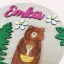 Dievčenská okrúhla menovka s medveďom, ihličnatými stromami, kvietkami a menom.