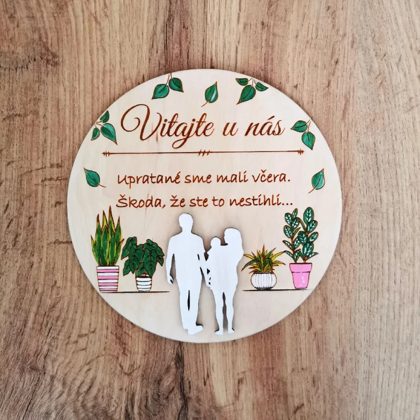 Drevená okrúhla tabuľka s textom "Vitajte u nás, upratané sme mali včera, škoda že ste to nestihli", so siluetami postáv a s ručne maľovanými izbovými rastlinami. 