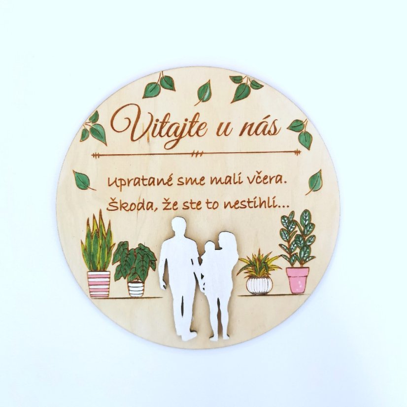 Drevená okrúhla tabuľka s textom "Vitajte u nás, upratané sme mali včera, škoda že ste to nestihli", so siluetami postáv a s ručne maľovanými izbovými rastlinami. 